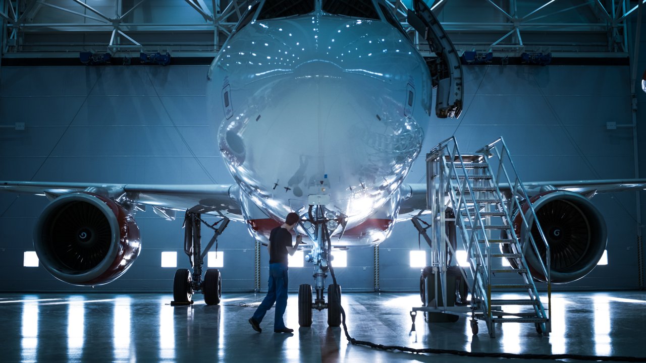 A large commercial aircraft is parked inside a spacious hangar, with a technician inspecting its landing gear