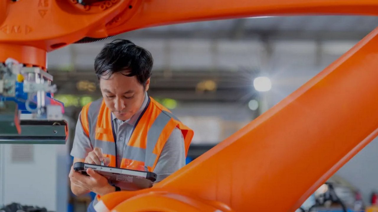 A worker wearing a high-visibility orange vest is using a tablet to inspect a large orange robotic arm in an industrial factory setting. 
