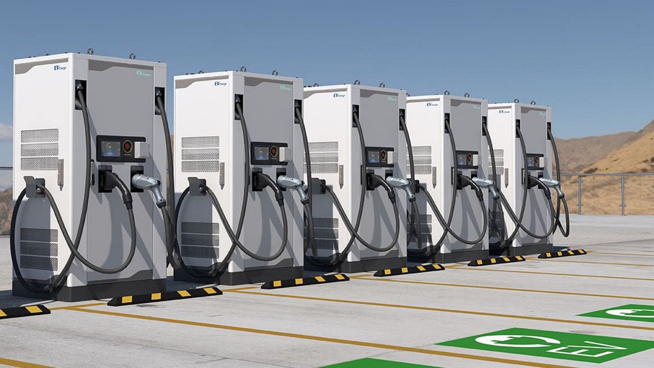 A row of modern electric vehicle charging stations is arranged in an outdoor parking lot under a clear blue sky. 