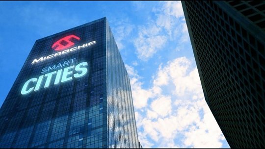 近代的な高層ビルのガラス外壁には「マイクロチップ スマートシティーズ」という文字が目立つように表示されています。建物は青空と散在する雲を背景にしており、都市革新を強調しています。