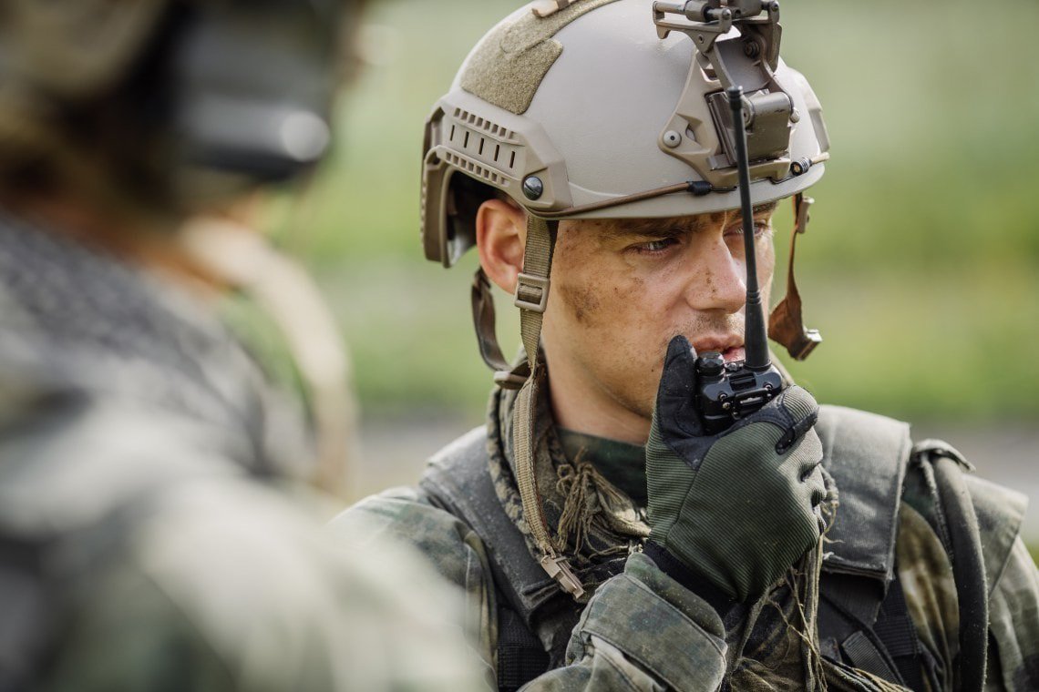 A soldier wearing advanced tactical gear and a helmet communicates using a handheld device in an outdoor setting.