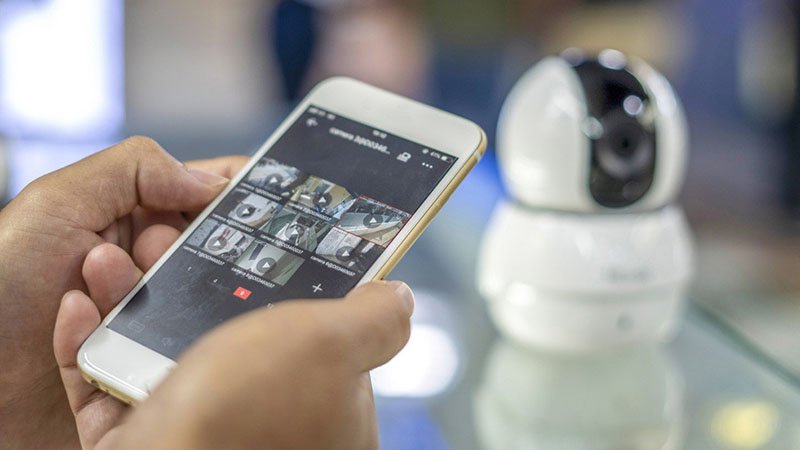 A person holds a smartphone displaying multiple camera feeds, suggesting remote monitoring capabilities.