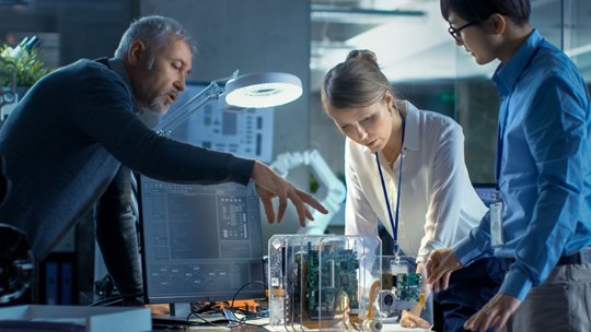 A group of professionals is working together in a modern office setting, focusing on an electronic prototype encased in a transparent housing