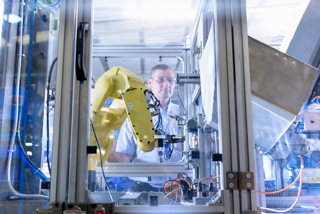 A yellow robotic arm operates within a glass-enclosed manufacturing environment. The setting features industrial machinery and wiring, indicating an automated production process. The scene is brightly lit, emphasizing the modern technology and precision engineering involved.