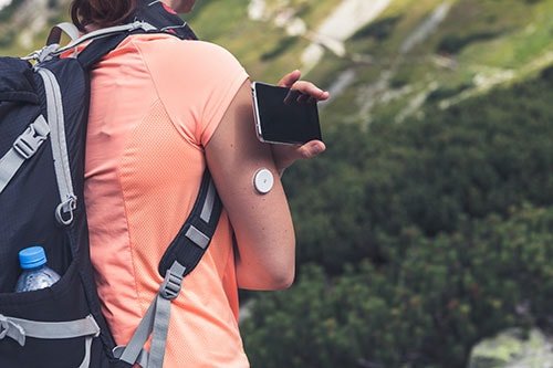 A woman wearing a peach-colored athletic shirt and a backpack is hiking in a mountainous outdoor setting. She has a wearable sensor attached to her arm and is holding a smartphone. The background features lush greenery and rugged terrain, suggesting an active and adventurous mood.