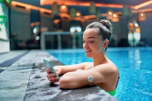 A woman leans on the edge of an indoor swimming pool while holding a smartphone. She is wearing a green swimsuit and has a small circular device attached to her upper arm. The setting features modern architecture with warm lighting and reflective water surfaces.
