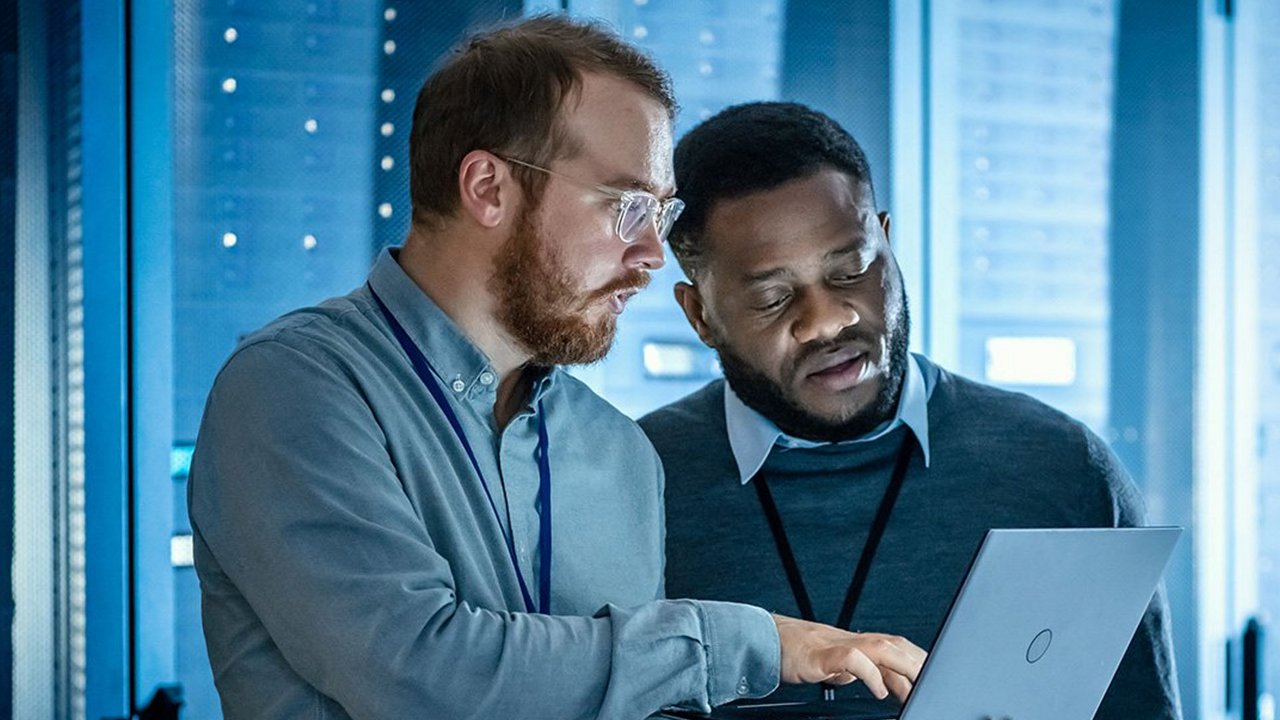Two individuals are working together on a laptop in a modern server room. 