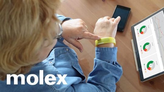 An older woman interacts with a yellow wearable device on her wrist while seated at a wooden table