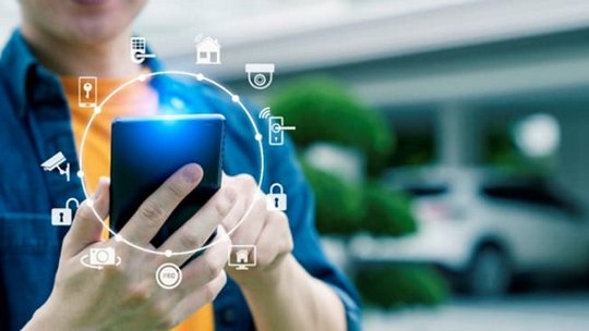 A close-up of a person holding a smartphone, interacting with smart home icons displayed digitally around the device.