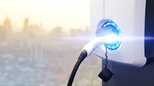 Un primo piano di una stazione di ricarica per veicoli elettrici con una luce blu luminosa che indica la ricarica in corso. Il cavo di ricarica è collegato saldamente alla stazione. Sullo sfondo si intravede uno skyline urbano sfocato, suggerendo un ambiente cittadino. L'immagine mette in evidenza energia pulita e tecnologia moderna.