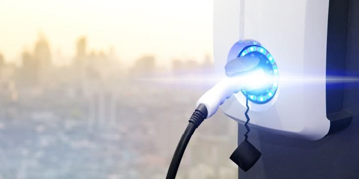 電気自動車充電ステーションのクローズアップビューで、充電が稼働中であることを示す青く光るランプが輝いています。充電ケーブルはステーションにしっかりと接続されています。背景にはぼやけた都会のスカイラインが映り込み、都市環境を示唆しています。この画像は、クリーンエネルギーと近代的な技術を際立たせています。