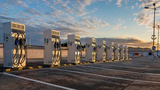 A row of modern electric vehicle charging stations is set in an open parking lot under a vibrant sunset sky