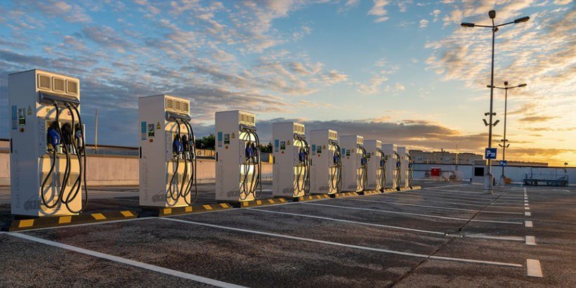 A row of modern electric vehicle charging stations is set in an open parking lot under a vibrant sunset sky