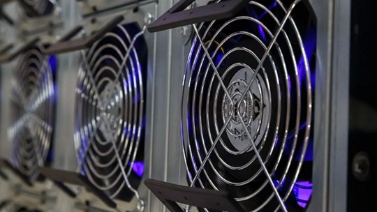 複数の産業用冷却ファンが一列に配置されている詳細な様子。ファンには金属製の格子が取り付けられており、稼働中であることを示すかのように淡い青紫色の光を放っています。この場所はデータセンターまたはマイニングリグ環境であると思われます。