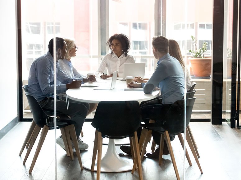 A group of professionals gathered around a round table in a glass-walled office. 