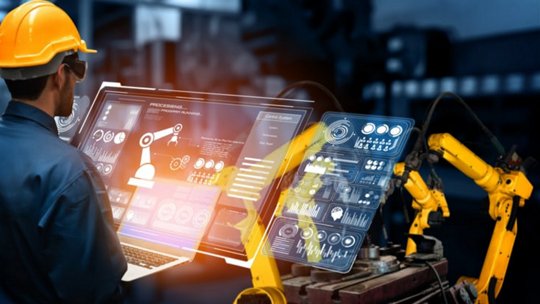 A male engineer wearing a yellow safety helmet operates a digital interface to control robotic arms in an industrial setting. 
