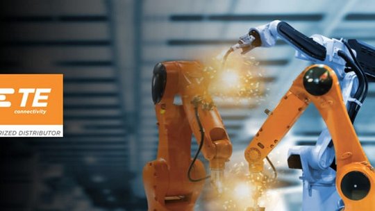 Two industrial robotic arms, one orange and one white, are shown in a factory setting performing tasks with visible sparks. 