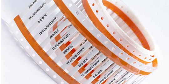 Rolls of orange and white TE Connectivity label tape with printed product information.