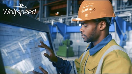 A worker wearing a safety helmet and high-visibility jacket interacts with a touchscreen in a modern industrial environment. 