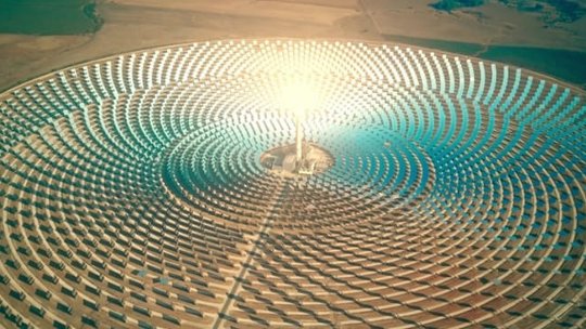 A large solar power plant featuring a central radiant tower surrounded by concentric arrays of mirrors. The setting appears to be a remote desert landscape under clear skies. The image highlights renewable energy technology and the geometric symmetry of the installation.