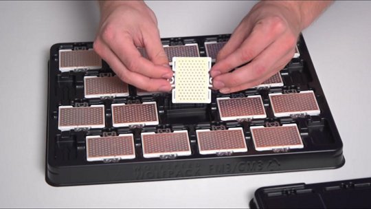 Close-up of hands holding a small electronic circuit board with a grid of components. The board is part of a larger tray containing multiple similar boards, arranged neatly. The setting appears to be a workspace or assembly area, emphasizing precision and organization.