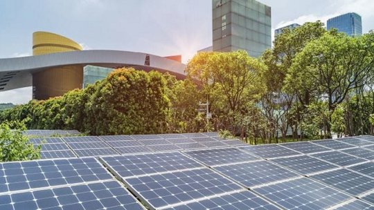A row of solar panels installed in a lush green area with trees and modern architecture in the background. 