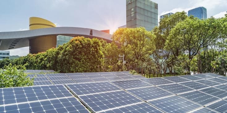 A row of solar panels installed in a lush green area with trees and modern architecture in the background. 
